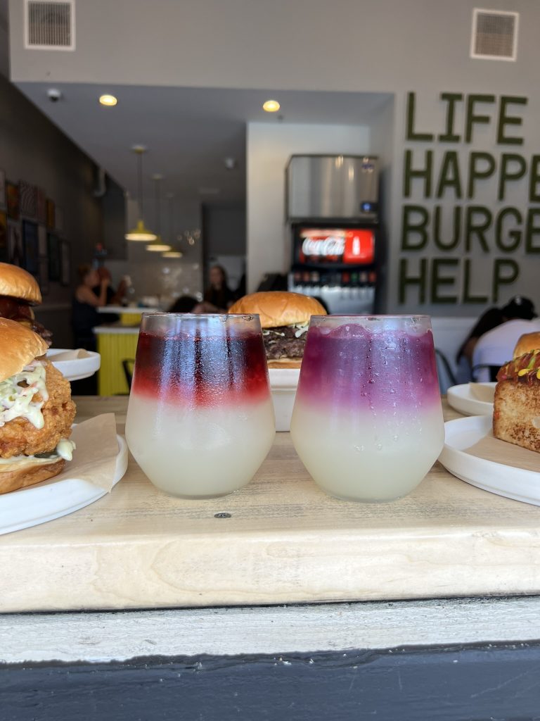 Two gradient drinks with a dark red top and light bottom, surrounded by burgers inside a restaurant.