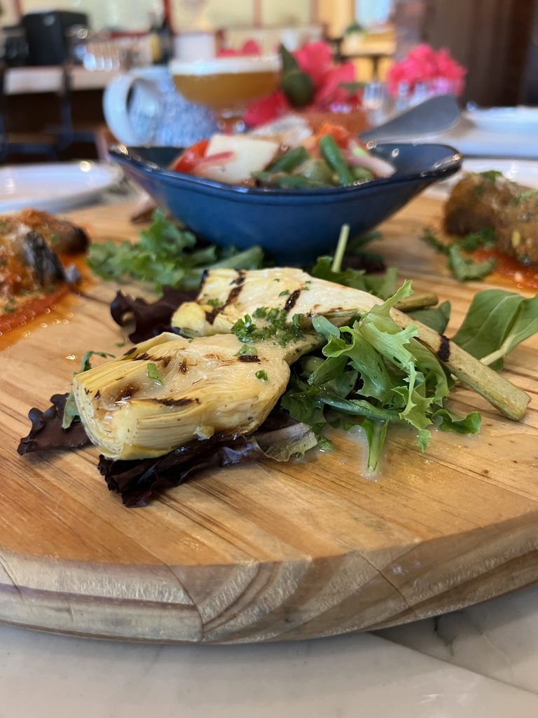 A wooden platter with grilled artichokes and fresh greens, accompanied by a bowl of mixed salad in the background.