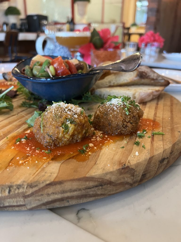 Two arancini on a wooden board with a bowl of mixed salad and slices of bread in the background.