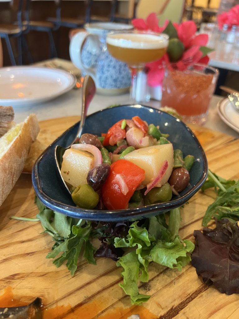 A colorful salad with olives, cherry tomatoes, and greens in a small blue bowl on a wooden board, surrounded by slices of bread and various drinks in the background.