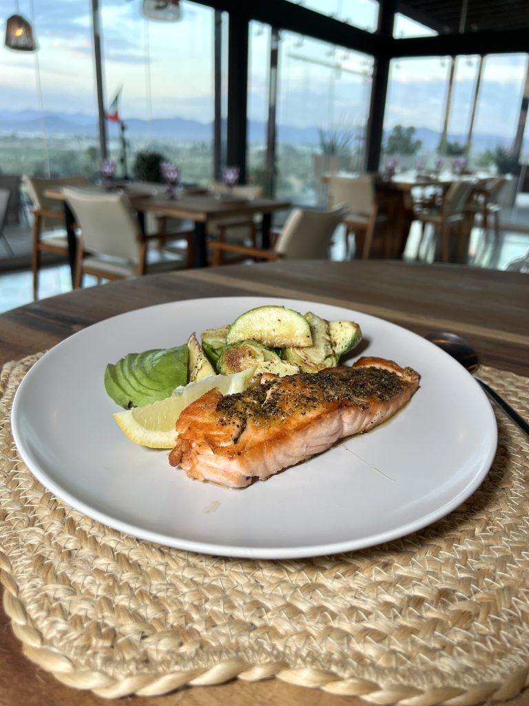 A plate with grilled salmon, lemon wedge, sliced avocado, and zucchini, set on a wooden table with a scenic view of a restaurant's glass walls and outdoor landscape.