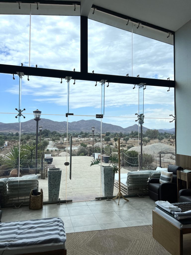 Modern interior with large glass windows overlooking a desert landscape and mountains, featuring tiled floors, lounge chairs, and potted plants.