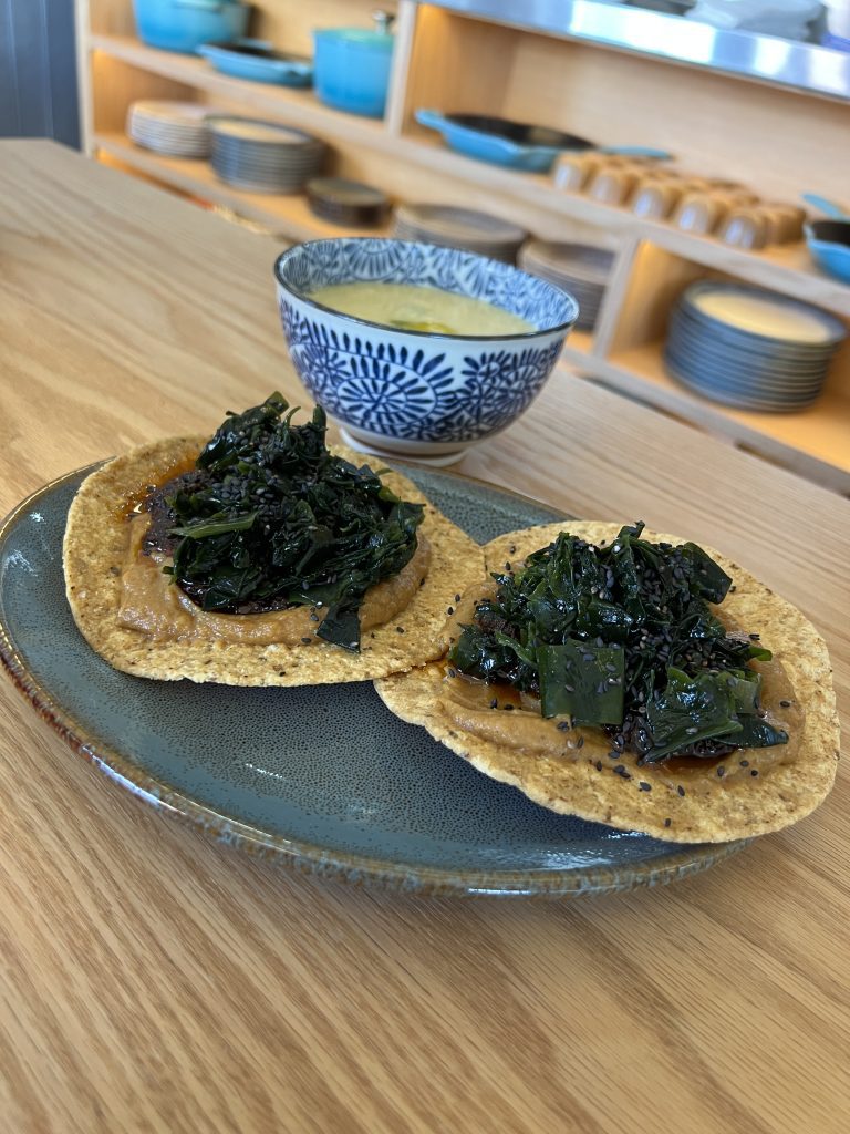 Two tostadas topped with greens on a plate, with a patterned bowl of soup in the background.