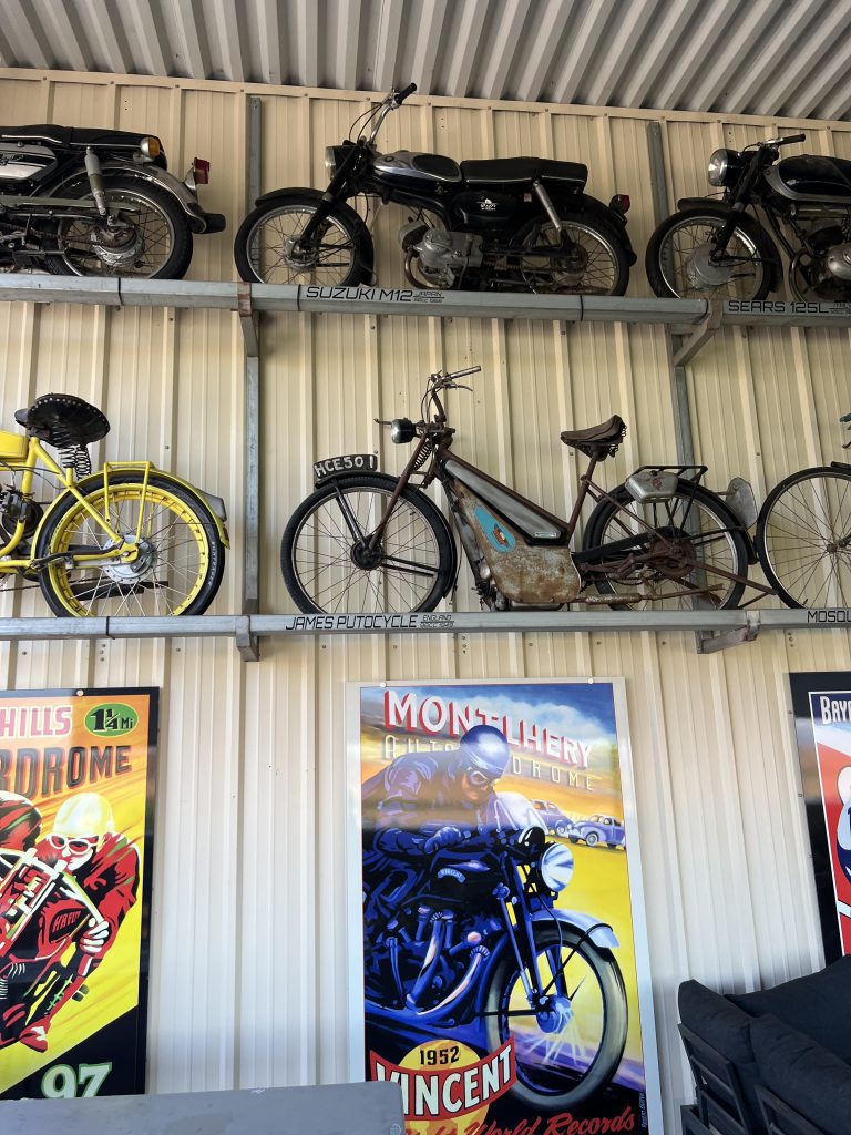 Vintage motorcycles and bicycles displayed on shelves against a corrugated wall, with vintage racing posters below.