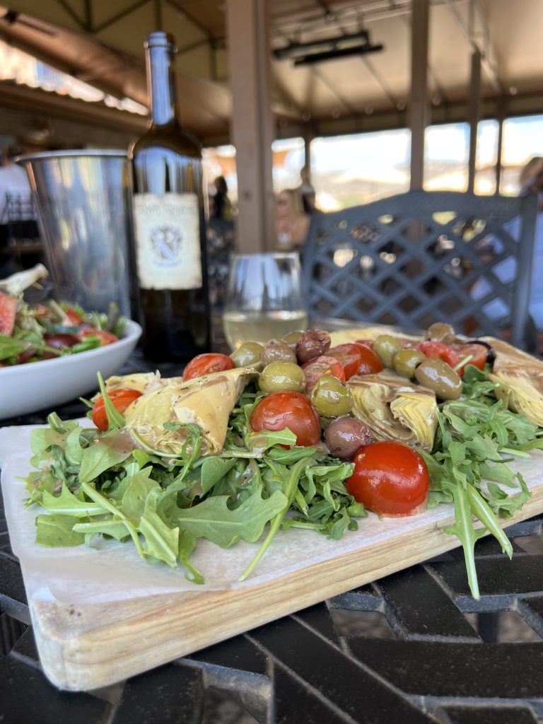 A salad with arugula, cherry tomatoes, olives, and artichoke hearts on a table, with a bottle of wine and glass in the background.