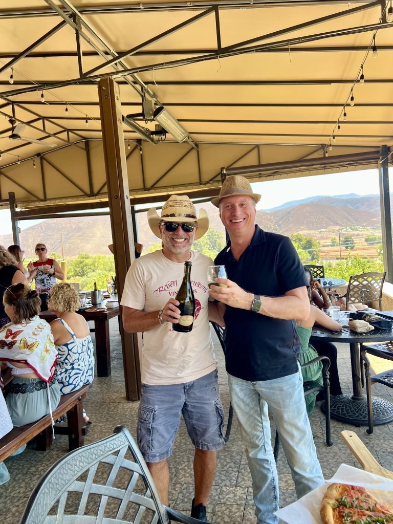 Two men wearing hats posing together under a patio roof, holding a bottle and glasses, with outdoor seating and mountains in the background.