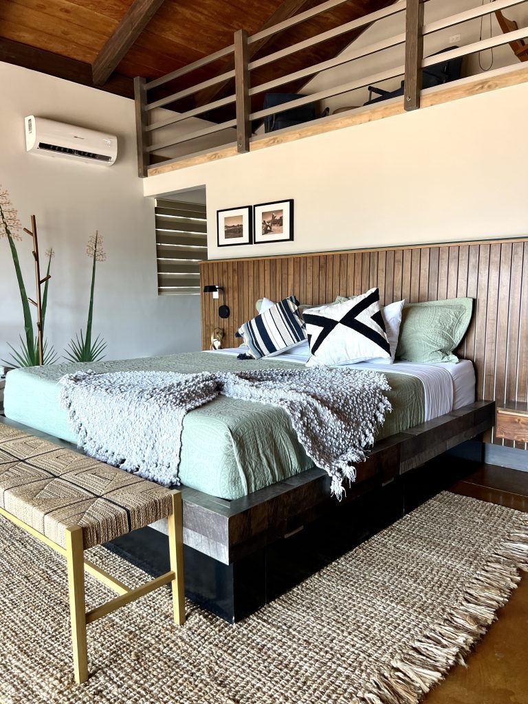 Modern bedroom with wooden accents, featuring a loft, green bedding, decorative pillows, and a woven bench on a textured rug.