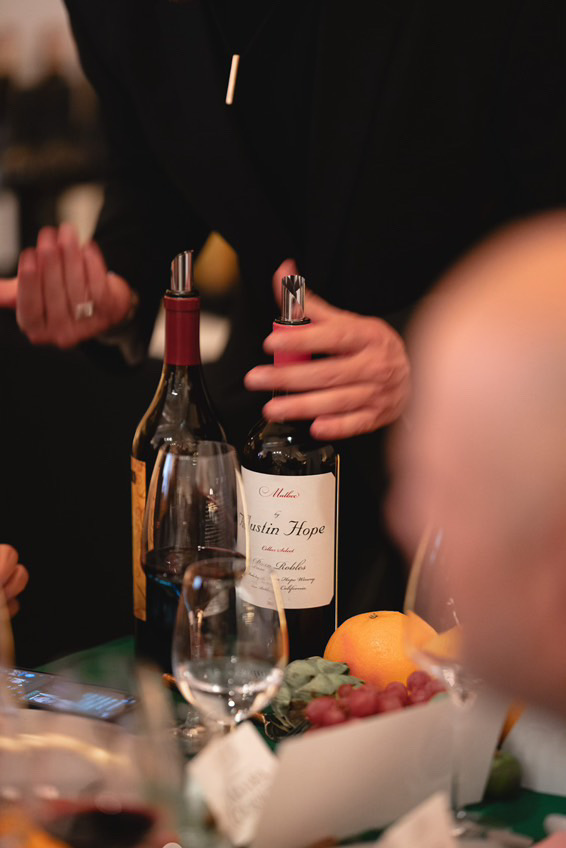A person holding a wine bottle labeled "Austin Hope" with glasses and fruit on the table.