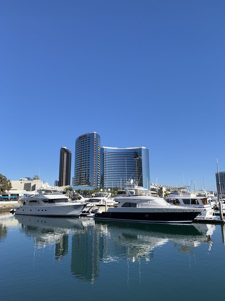 Yachts docked at a marina with modern buildings in the background, under a clear blue sky.