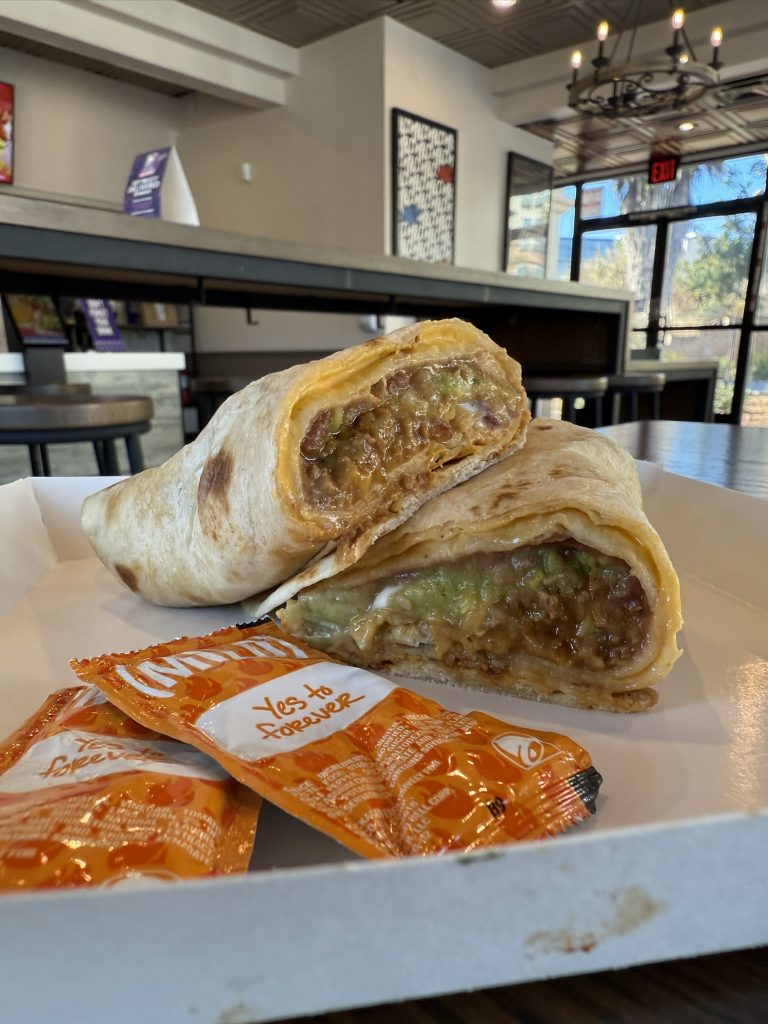 Cut burrito with visible fillings on a plate, accompanied by two hot sauce packets on a table in a restaurant.