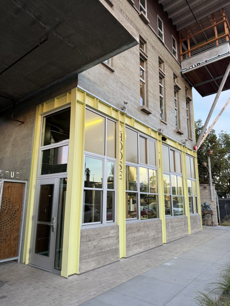 Tall beige building with yellow accents and large windows on the ground floor.