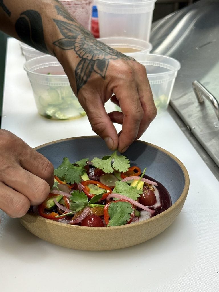 Tattooed hands garnishing a dish with fresh cilantro in a kitchen.