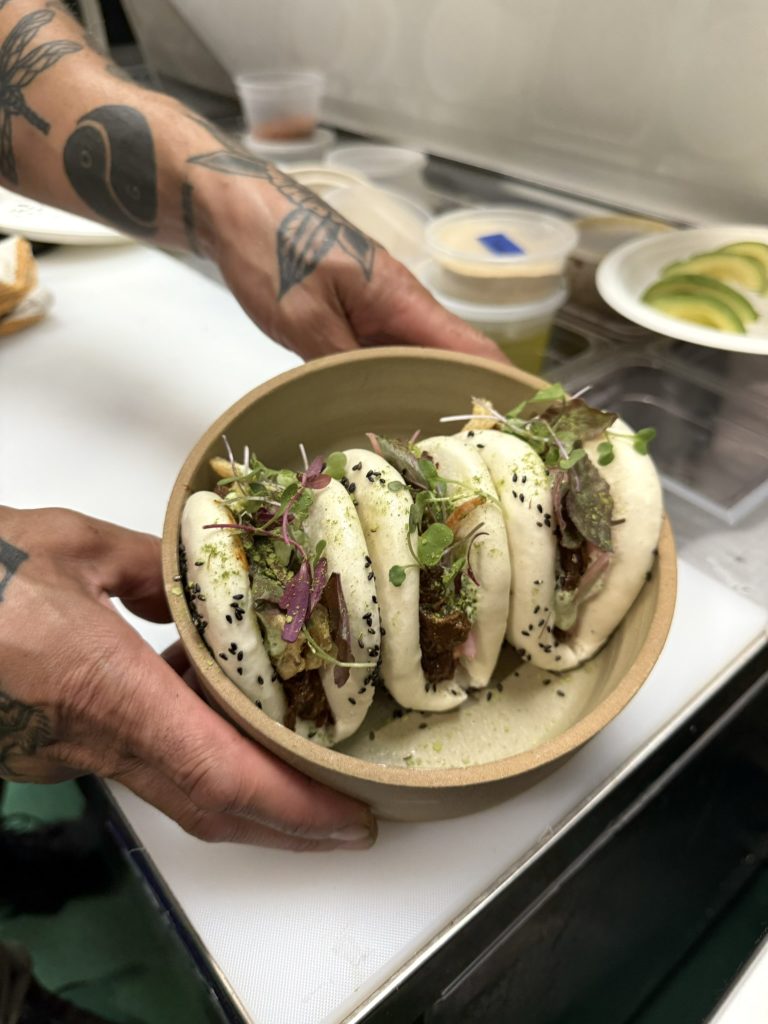 A person holding a bowl of three steamed bao buns filled with greens and garnished with black sesame seeds.