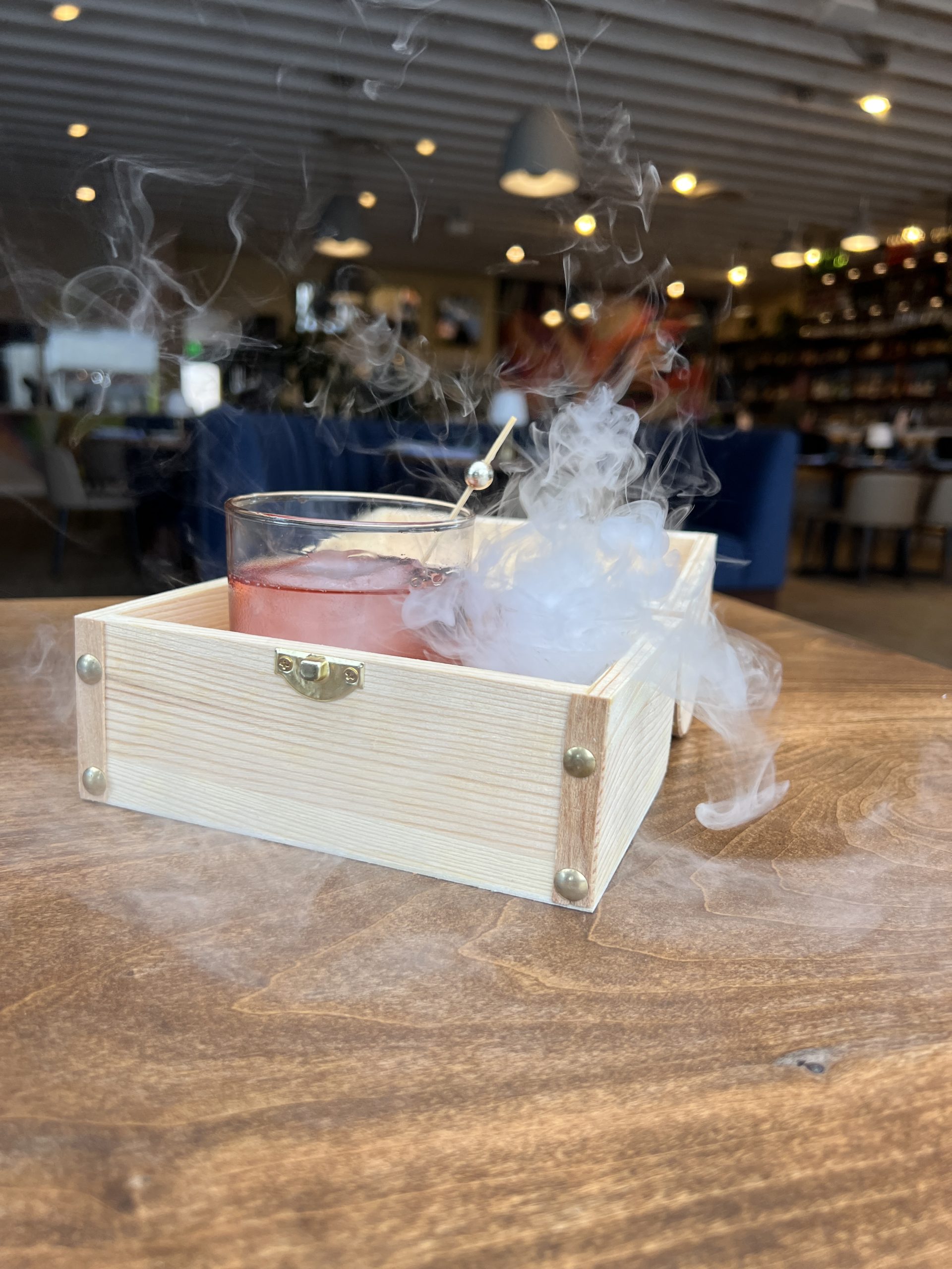 A cocktail in a glass resting in a wooden box with smoke swirling around it on a wooden table.