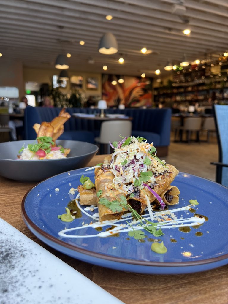 Rolled tacos topped with shredded cabbage, herbs, and sauce on a blue plate in a restaurant setting.