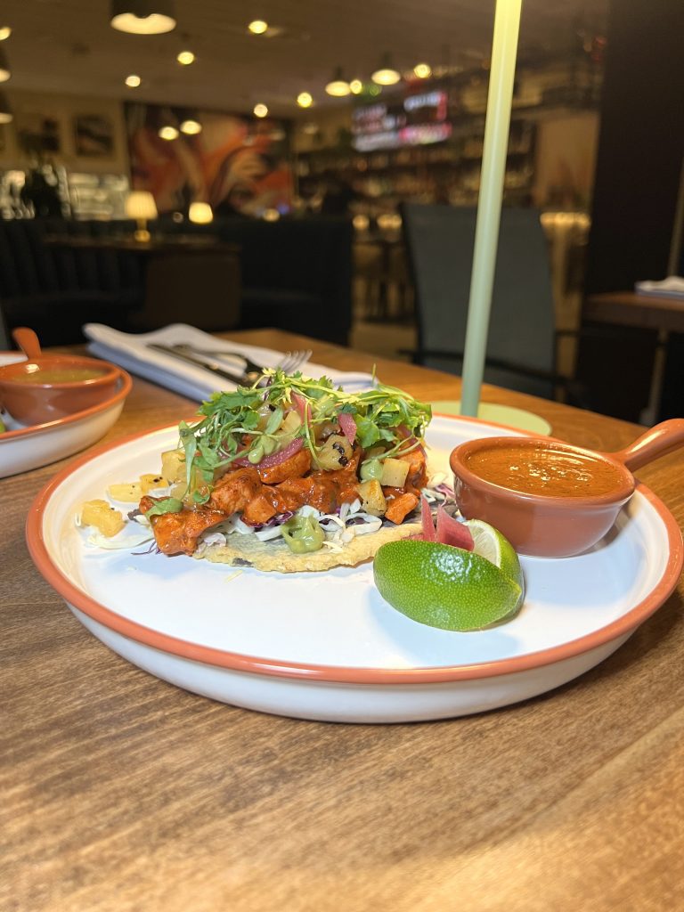 A plate with a taco topped with meat, vegetables, and greens, served with lime and a small dish of sauce.