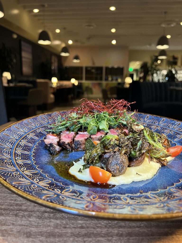 A decorative plate with seared steak slices, greens, cherry tomatoes, and grilled vegetables on a bed of puree in an elegant dining setting.