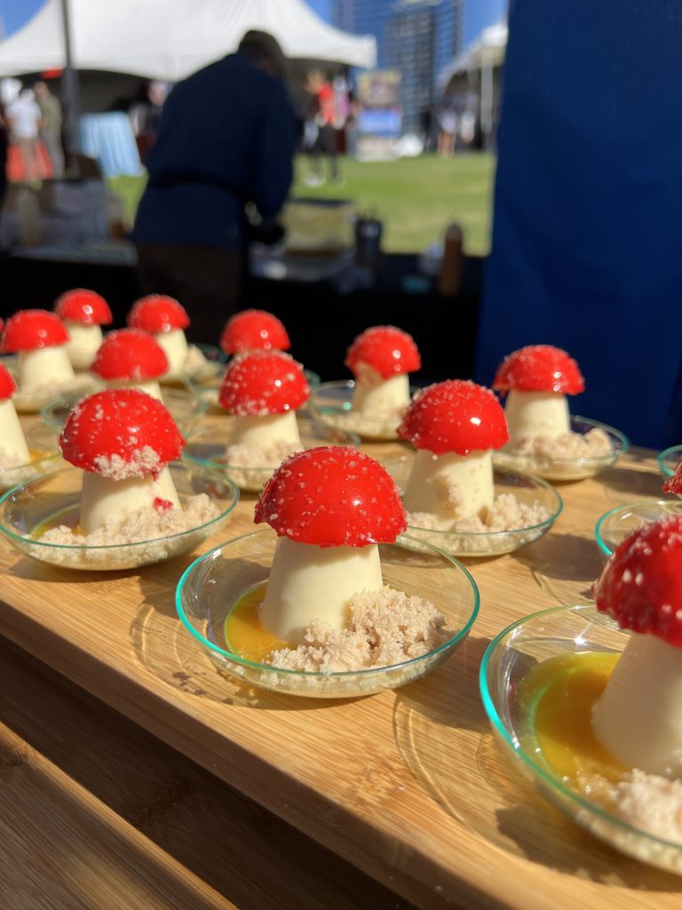 Miniature mushroom-shaped desserts with red tops on small glass plates, displayed on a wooden board at an outdoor event.