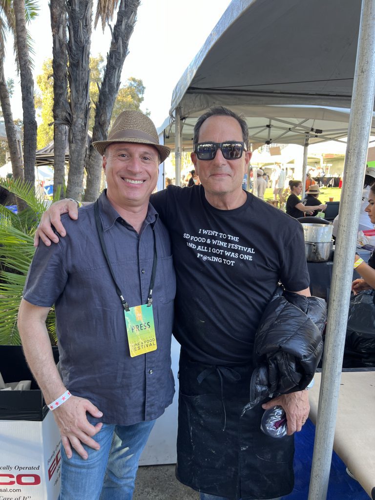 Two men smiling at the camera under a tent at an outdoor event, with one wearing a straw hat and press badge.