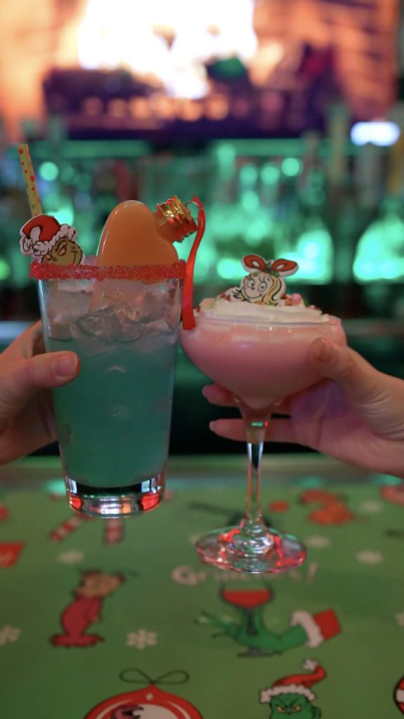 Two festive cocktails, one blue with an orange garnish and one pink with whipped cream, adorned with holiday-themed decorations, being held up in a cheers gesture.