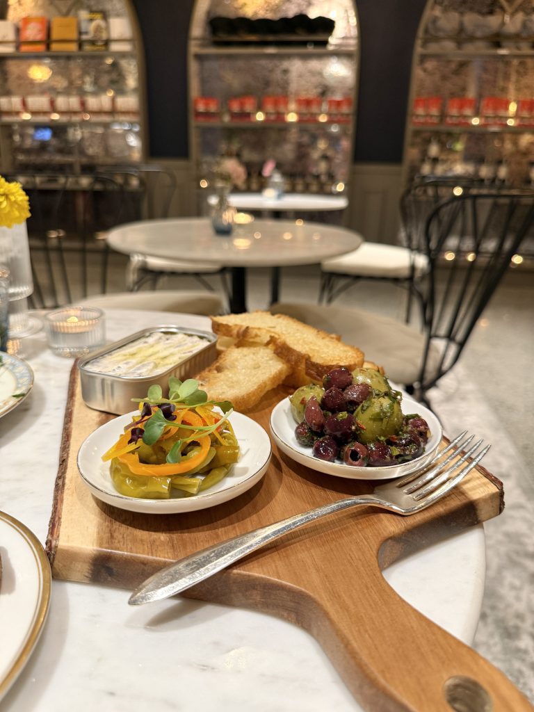 A wooden serving board with small dishes of pickled vegetables and olives, alongside toasted bread slices, on a marble table in an elegantly decorated restaurant.