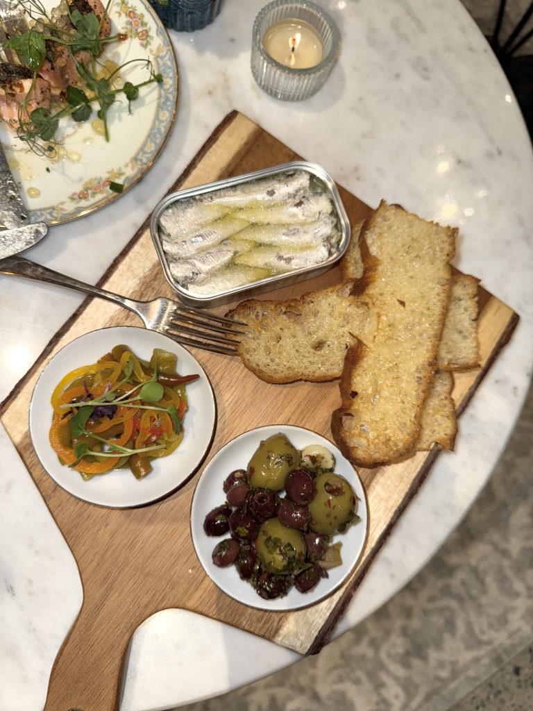 A wooden serving board with sardines, toasted bread slices, a dish of olives, and a dish of marinated peppers on a marble table.