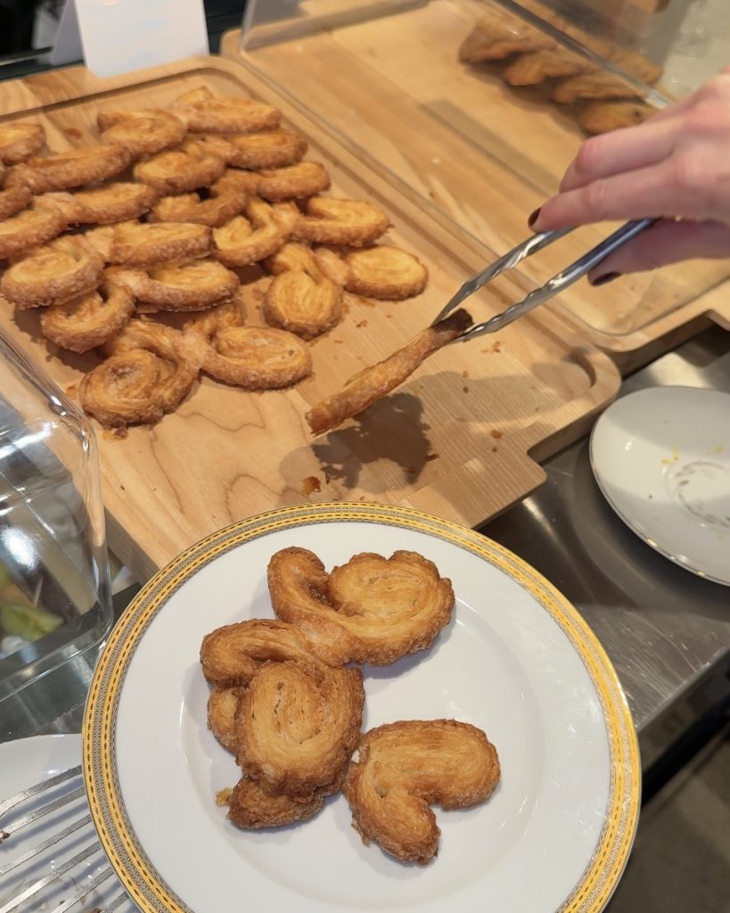 A person uses tongs to pick up palmiers from a wooden board, placing them on a decorative plate.