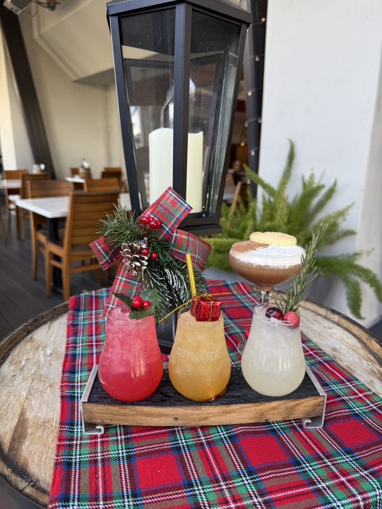 Three festive cocktails garnished with holiday decorations on a wooden tray, set on a plaid tablecloth, with a lantern and wooden chairs in the background.