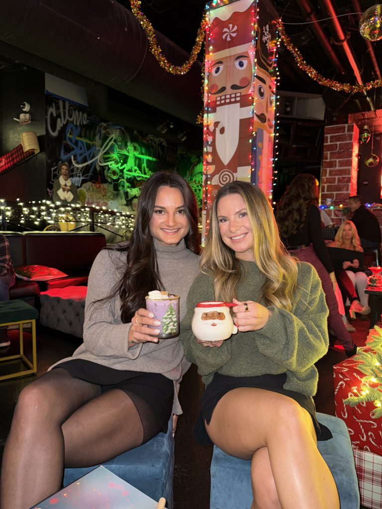 Two women sitting indoors with festive holiday decorations, holding Christmas-themed mugs and smiling.