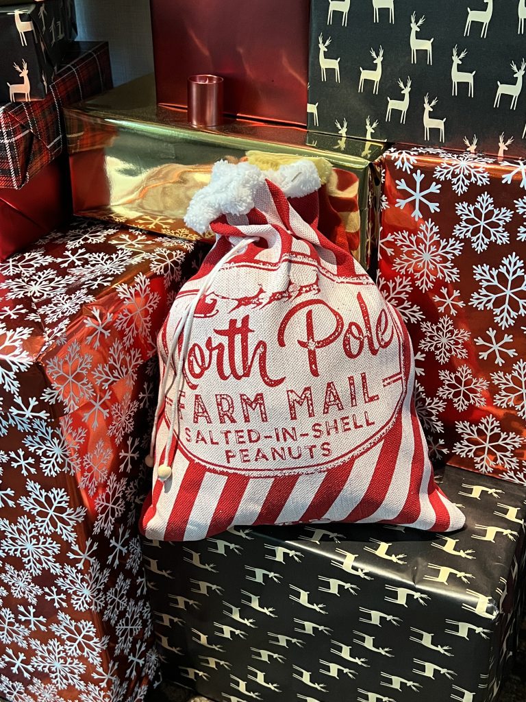 A sack labeled "North Pole Farm Mail Salted-In-Shell Peanuts" in red and white stripes, surrounded by gift boxes with snowflake, reindeer, and plaid patterns.