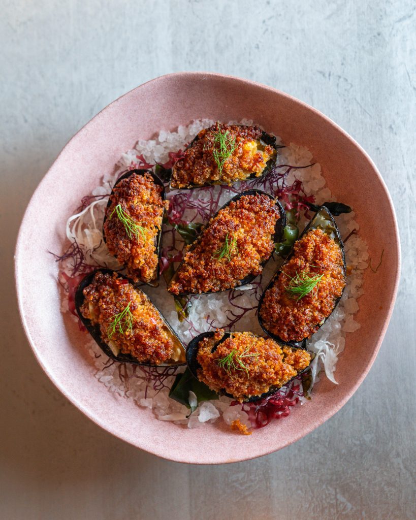 A bowl of baked mussels topped with breadcrumbs and garnished with herbs on a bed of seaweed and rock salt.