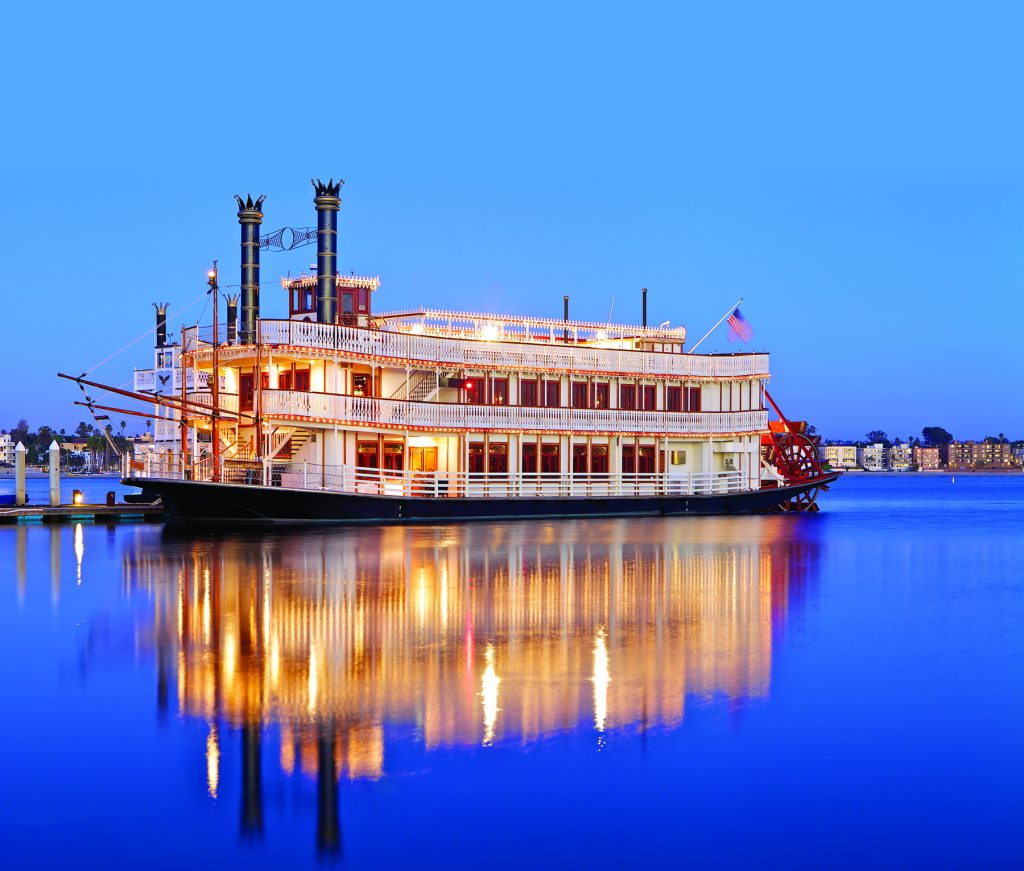 A brightly lit riverboat docked on calm blue water, reflecting its lights, with a clear evening sky.