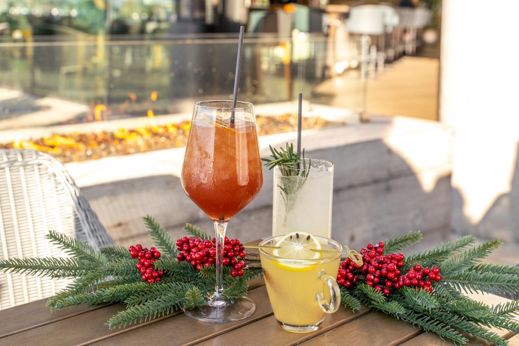 Festive drinks on a table with holiday decorations, featuring a cocktail in a wine glass, a drink in a tall glass with rosemary, and a yellow beverage in a clear mug garnished with a lemon slice.