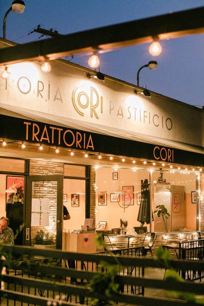Outdoor evening view of a warmly lit restaurant named "Trattoria Cori" with string lights and a few tables visible inside.